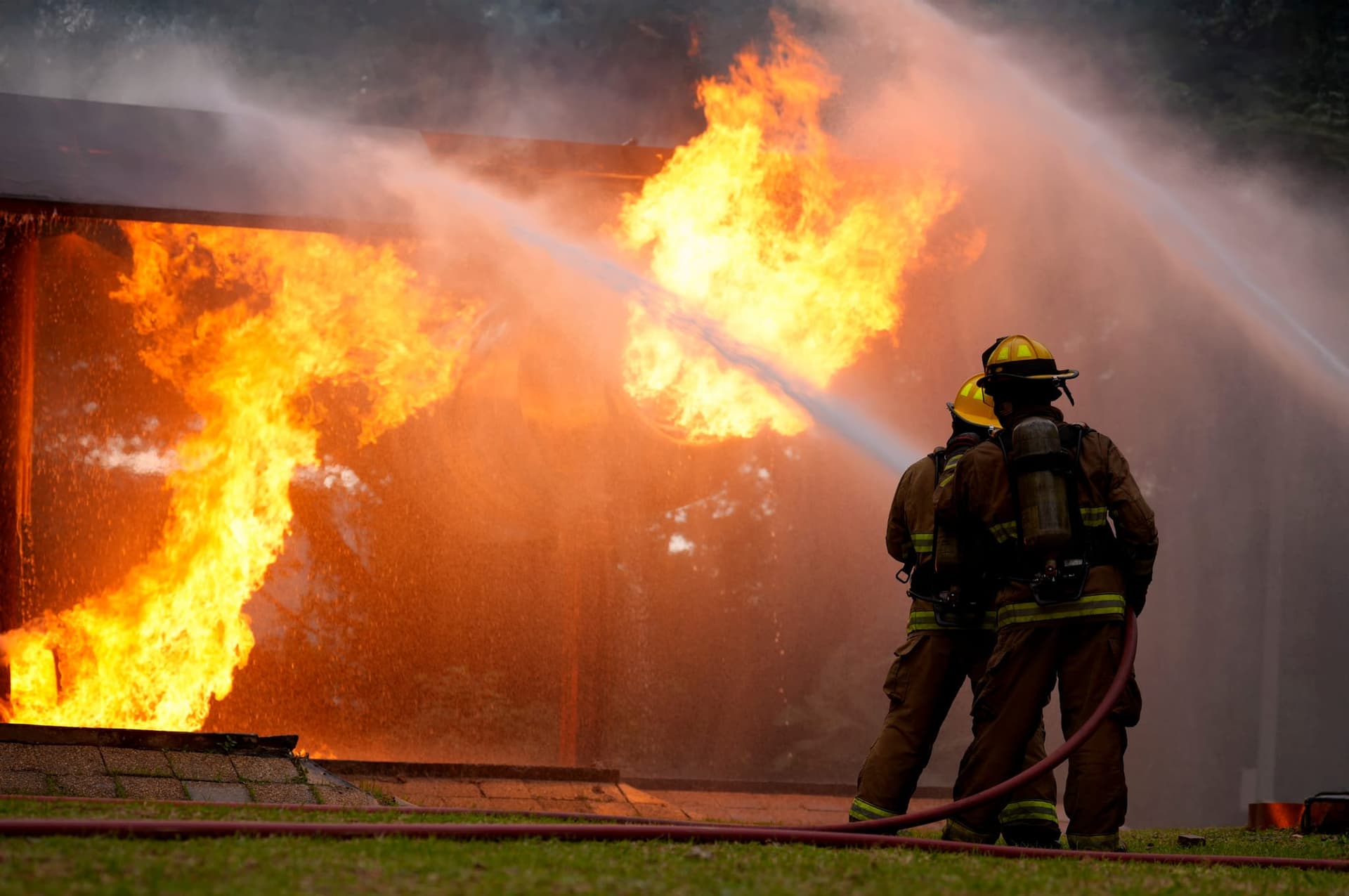 Two firefighters in full turnout gear directing a hose stream into flames pouring from the side of a building