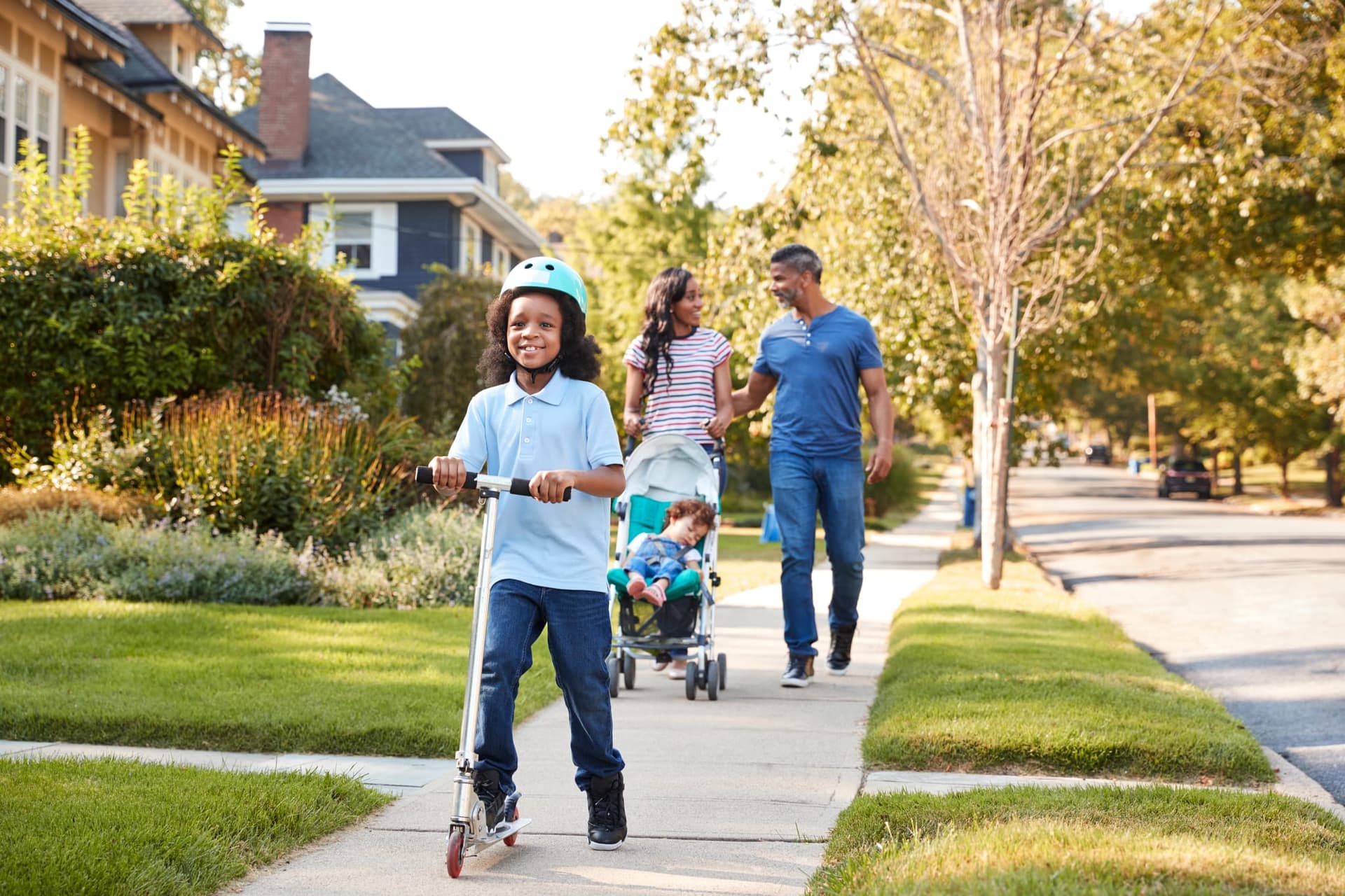 A family walking a tree-lined residential sidewalk, a child riding a scooter ahead of adults pushing a stroller