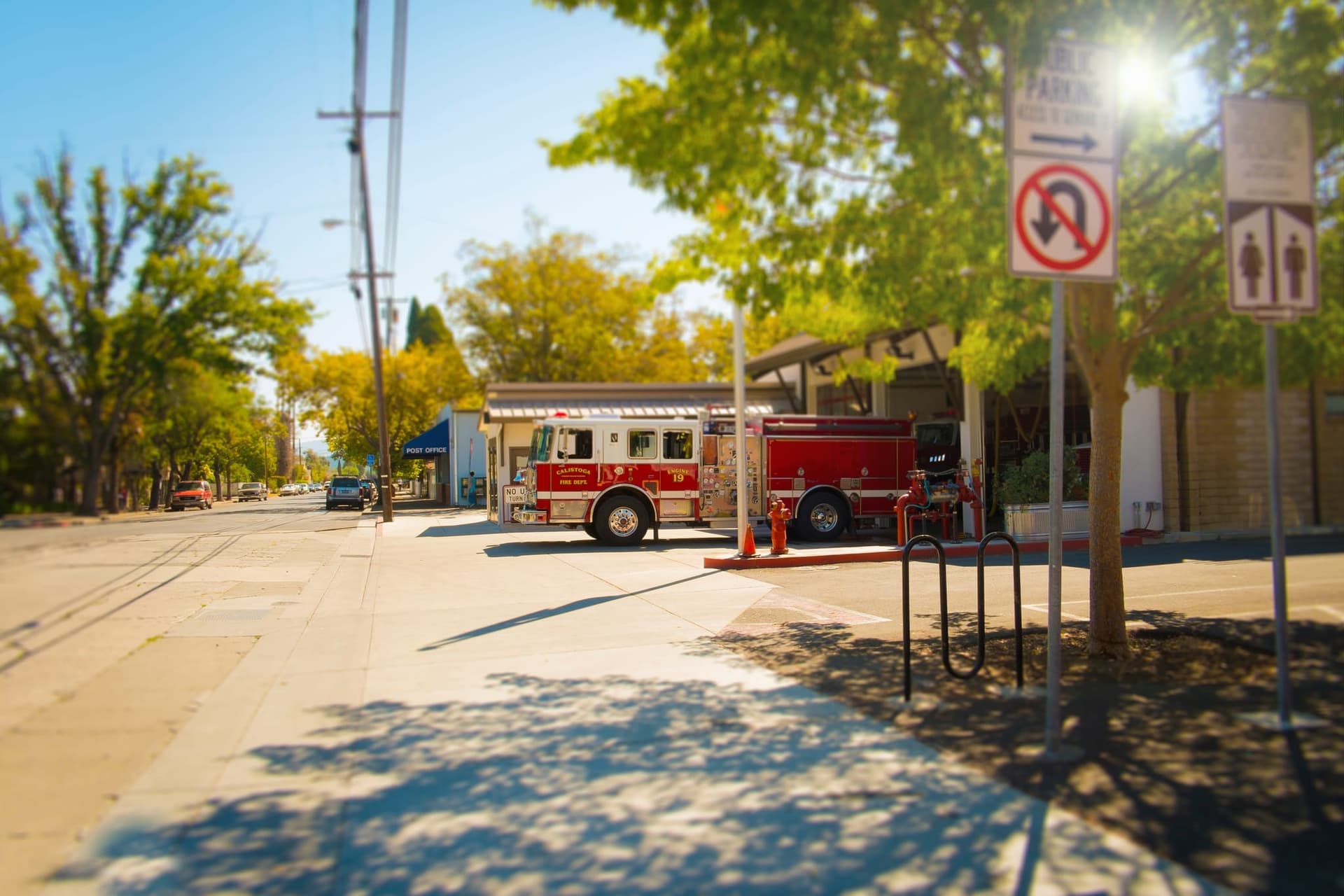A fire engine pulling out of a small-town fire station onto a tree-lined street, with a post office and parking signs nearby